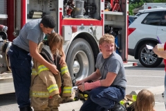 Firefighters help a child try on firefighting gear beside a fire truck during Cape Coral Kiwanis KidsFest 2025.