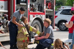Child wearing oversized firefighter turnout gear receives assistance from firefighters at Cape Coral Kiwanis KidsFest 2025.