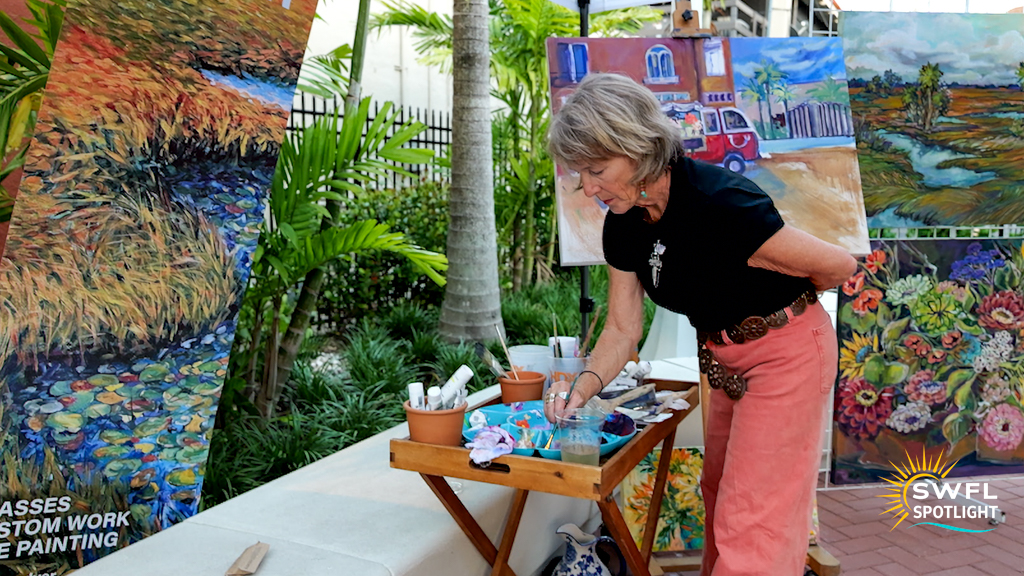 Artist Marie Dyer paints outdoors during Fort Myers Art Walk, surrounded by colorful landscape and floral artwork in the River District.
