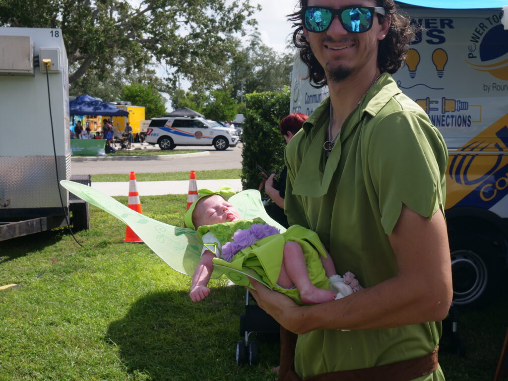 A father in a Peter Pan costume smiles while holding his newborn baby dressed as Tinkerbell during an outdoor family festival.