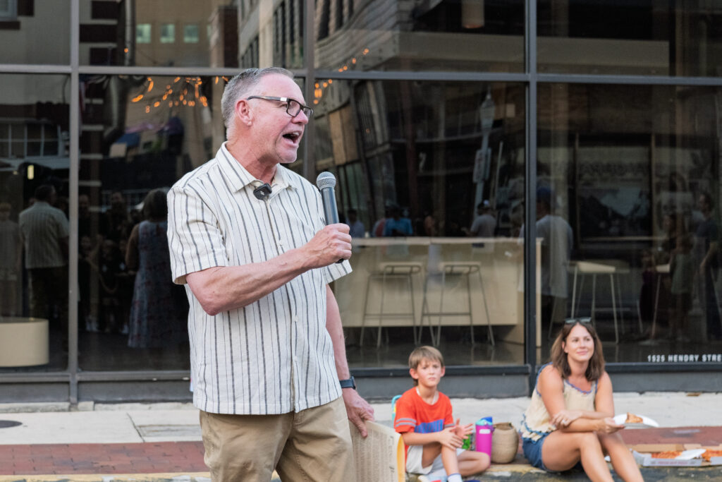 Fort Myers Mayor Kevin B. Anderson speaks into a microphone during a dedication for Downtown House of Pizza, with attendees seated nearby.